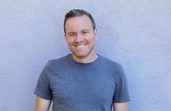 A smiling man with short brown hair wearing a grey t-shirt against a light blue textured wall.
