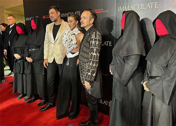People in formal attire and figures in black hooded robes with pink face coverings pose on a red carpet.