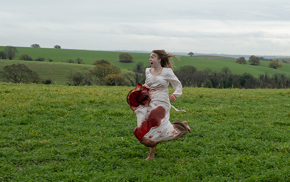 A woman in a white dress with large red stains runs through a vast green field under an overcast sky.