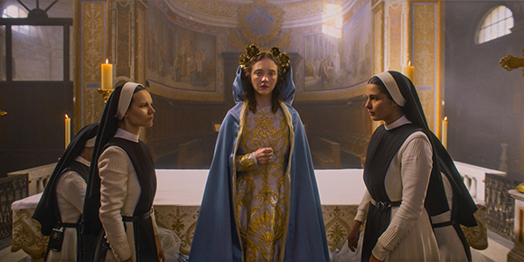 A woman in a blue and gold gown stands between three nuns in a grand, ornate hall.