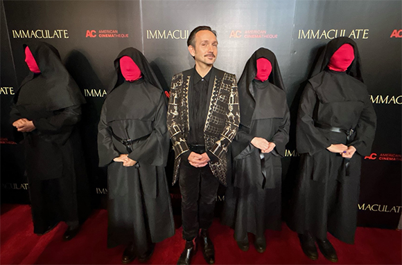 A man in a patterned blazer stands flanked by four figures in black robes with red face masks.