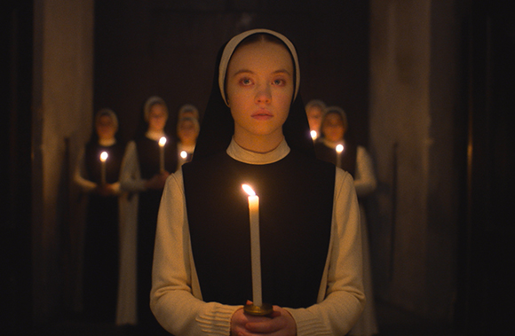 A young nun in a black and white habit holds a lit candle, leading a line of nuns in a dark corridor.
