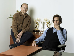 Two men sit in an office with two gold trophies on a wooden cabinet behind them.
