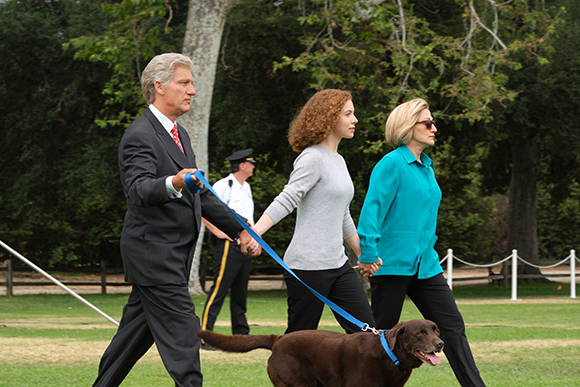 A man in a suit walks a brown dog on a blue leash, followed by two women walking side-by-side.