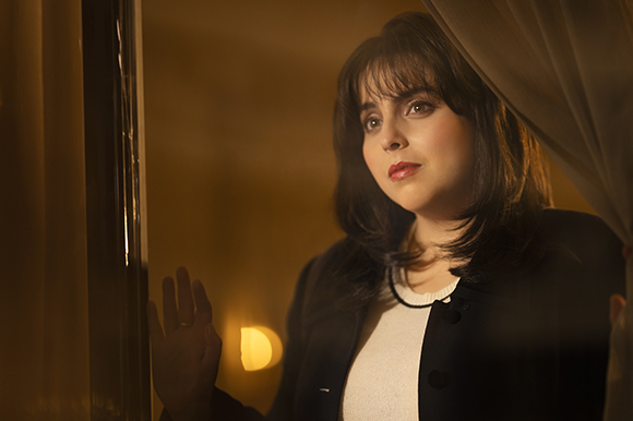 A woman with dark hair and red lipstick gazes thoughtfully through a window at night.