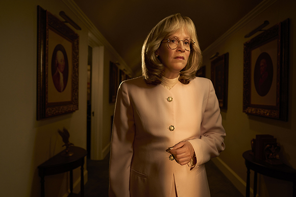 A woman with blonde hair and glasses wearing a pale pink blazer stands in a hallway with framed portraits.