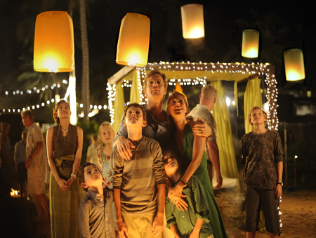 A family and group of people look upward at glowing yellow sky lanterns in a dark outdoor setting.