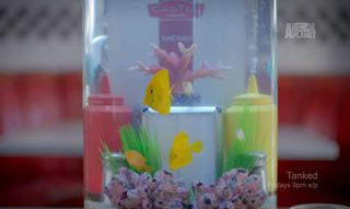 A small cylindrical fish tank with yellow fish, pink coral, and colorful condiment-shaped decorations.