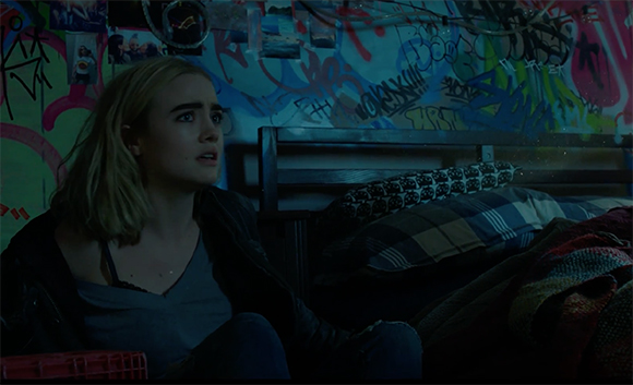 A blonde woman with a concerned expression sits in a bedroom with graffiti-covered walls.