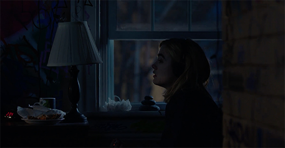 A person in profile looking out a dark window next to a lit lamp and a plate of food.