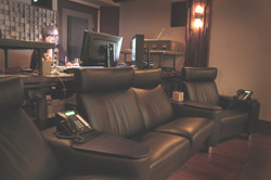A person works at a desk with monitors next to a black leather sofa and armchair.