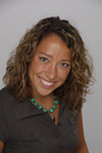 A smiling woman with curly brown hair and a turquoise beaded necklace against a gray background.