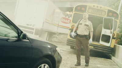 A man in a tan uniform stands in front of a yellow charter bus and a white truck in a dusty scene.