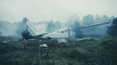 A white small aircraft sits in a grassy field amidst smoke and a backdrop of pine trees.