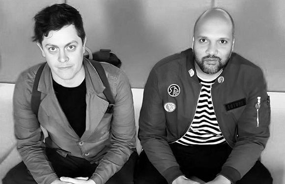 Two men sitting next to each other, looking directly at the camera in a black and white portrait.