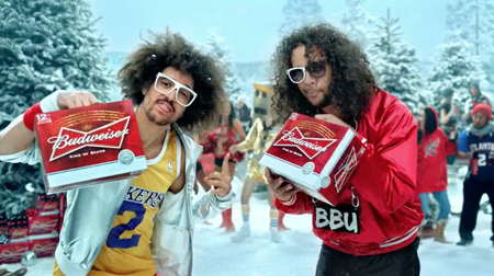 Two men with large curly hair and sunglasses hold Budweiser beer packs in a snowy outdoor landscape.