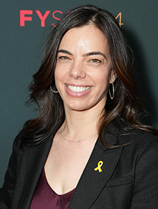A woman with dark hair smiling, wearing a black blazer and a small yellow ribbon pin.