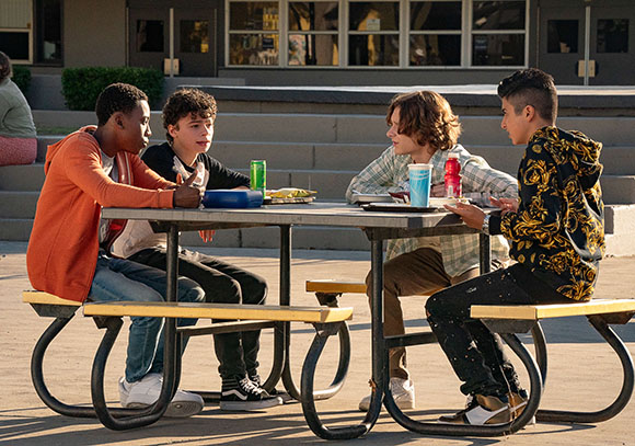 Four teenage boys sit around a picnic table outdoors, talking and eating.