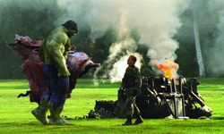 A large green humanoid figure stands in a field facing a man near a smoking, overturned vehicle.