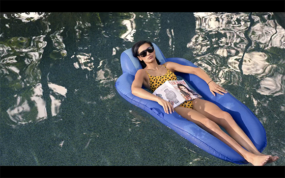 A woman in a yellow polka dot swimsuit relaxes on a blue inflatable float in a swimming pool.