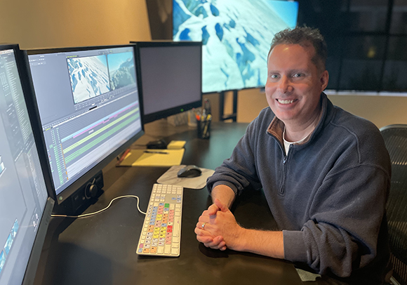 A smiling man sits at a desk with three computer monitors and a keyboard in a workspace.