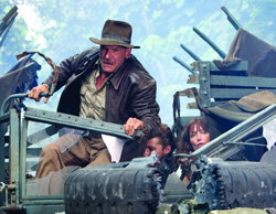 A man in a fedora and leather jacket leans out of a wrecked vehicle alongside two other people.