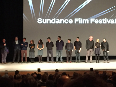 A group of people stand on a stage in front of a screen displaying the Sundance Film Festival logo.