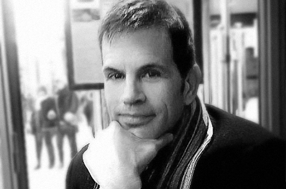 Black and white close-up portrait of a man looking at the camera with his hand under his chin.