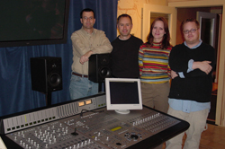 Four people stand behind a professional audio mixing console and monitor in a recording studio.