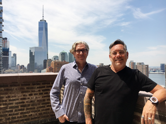 Two men smiling on a rooftop with a cityscape and One World Trade Center under a blue sky.
