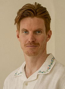 A man with reddish-brown hair and a mustache wearing a white embroidered shirt.