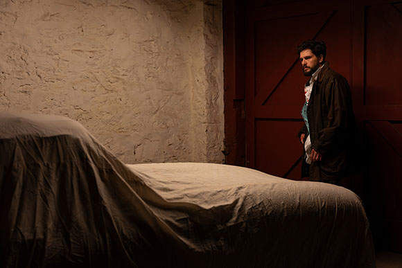 A man with dark hair and a beard stands in a dimly lit room next to a bed draped in a white sheet.