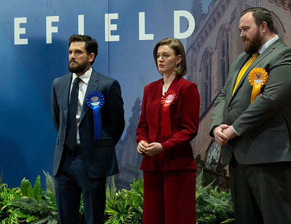 Three people in suits wearing colorful rosettes stand before a blue backdrop with white text.