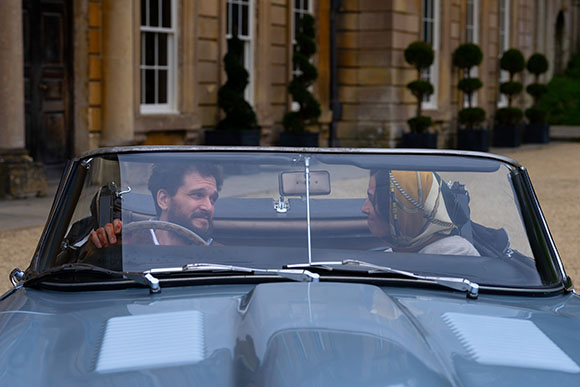A man and a woman sit in a vintage car, facing one another.