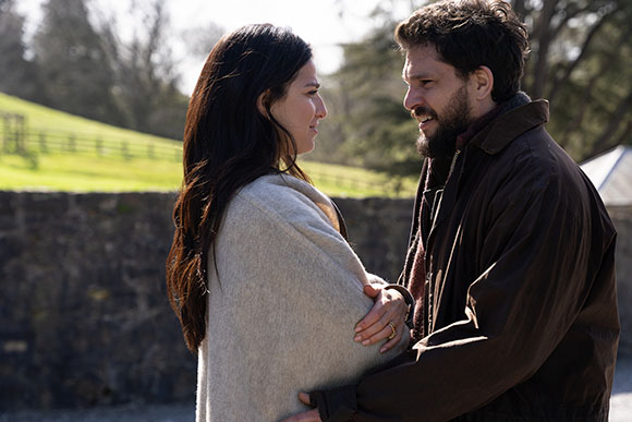 A man with a beard and a woman with long dark hair look at each other while standing close together.