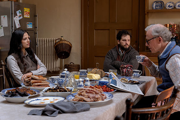 A woman and two men sit at a table filled with breakfast food, including eggs, bacon, and sausages.