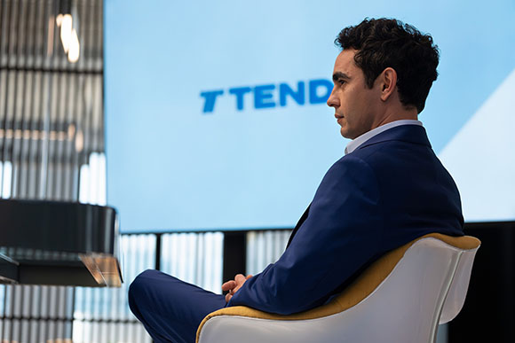 A man in a blue suit sits in a white chair, looking toward a large blue screen with the word TEND.