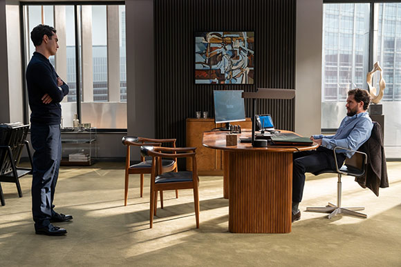 A man stands with arms crossed facing another man seated at a large wooden desk in a modern office.