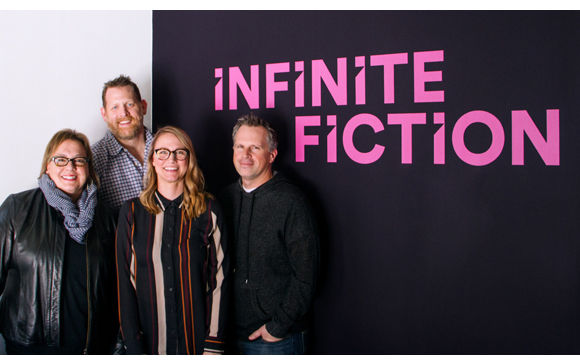 Four smiling adults pose in front of a large black wall with the pink text "INFINITE FICTION".