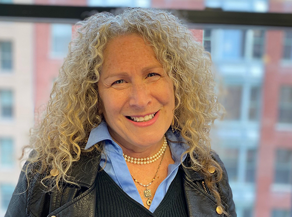 A smiling woman with long, curly blonde hair wearing a light blue shirt, black vest, and leather jacket.