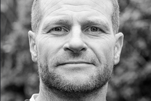 Black and white close-up portrait of a man with short hair and a beard looking directly at the camera.
