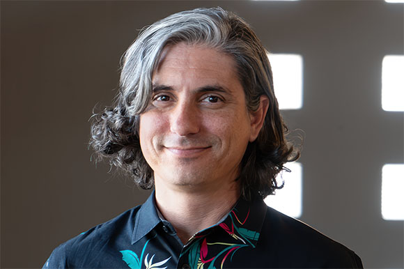 A man with long, wavy grey and black hair wearing a black floral shirt.