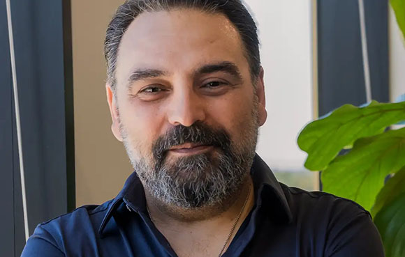 A man with dark hair and a salt-and-pepper beard smiling slightly in a dark shirt.