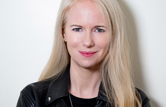 A blonde woman with blue eyes and a slight smile wearing a black leather jacket against a white background.