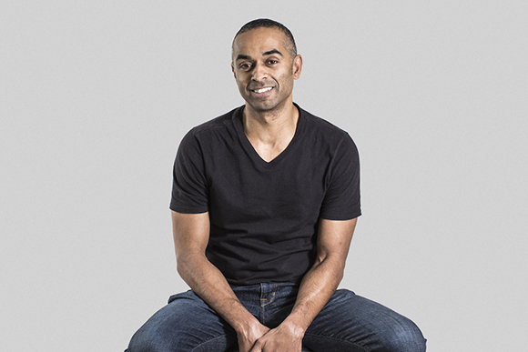 A smiling man with short dark hair wearing a black v-neck t-shirt and blue jeans, sitting against a gray backdrop.