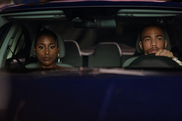 A man and a woman sit in the front seats of a car at night, looking forward with serious expressions.