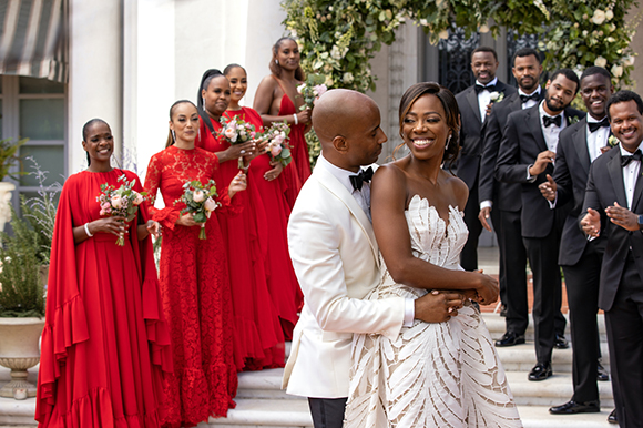 A bride and groom embrace in front of bridesmaids in red and groomsmen in black tuxedos.