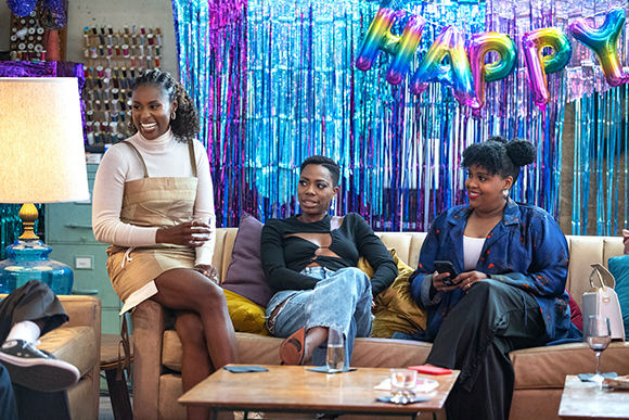 Three women sit on a tan sofa in front of blue and purple tinsel decorations and large colorful letters.