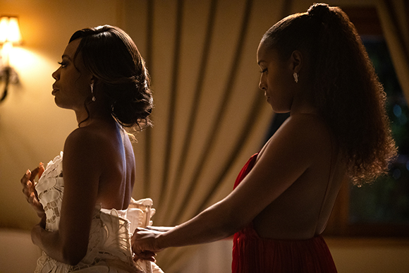 A woman in a white lace dress stands while another woman in a red dress adjusts the back of her garment.