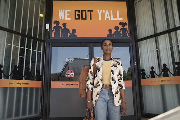 A woman in a patterned shirt and jeans stands before a building featuring a large orange sign with silhouettes.